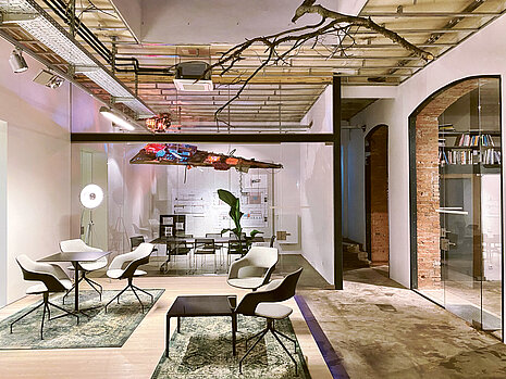 In a waiting area, Occo chairs with black seat shells and gray seat and back upholstery stand around small black tables. In the background, a meeting room can be seen behind a glass wall, where black Aline chairs are arranged around a table. The space is furnished in an industrial style.