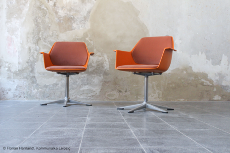 Here are two chairs in a warm orange hue. The chairs combine clean lines with a sturdy four-star metal base. Against the rough concrete wall, an intriguing contrast emerges between modern design and the industrial setting.
