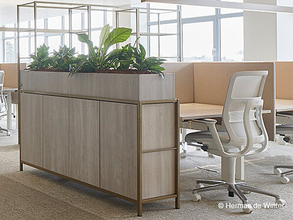 The photo shows a modern, bright open-plan office with plenty of natural light, wood finishes, and large plant displays used as room dividers. White AT swivel chairs can be seen at the workstations.