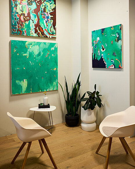 Two Yonda chairs with white seat shells, gray felt pads and wooden four-legged frames are in the foreground. In the background is a white wall with green pictures hanging on it. There are also plants and a side table.