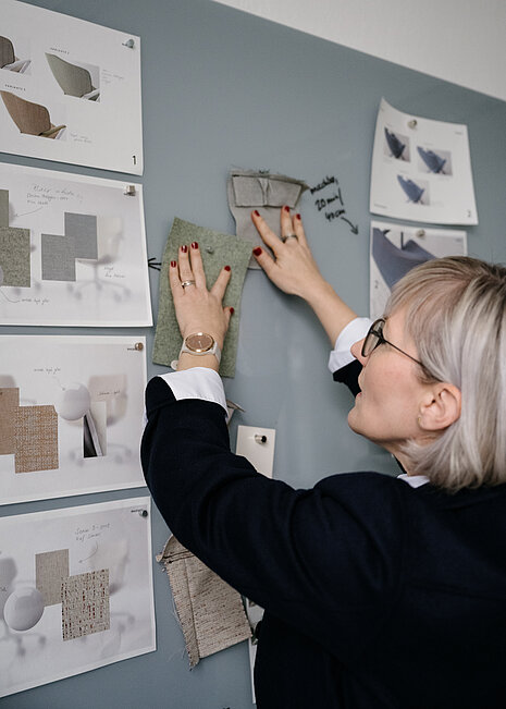 Our colleague Julia Wilkening-Martin is arranging fabric swatches and material collages on a magnetic board to select suitable upholstery and colors for an Intra chair. The sketches and mood boards in the background show the various stages of the design process.