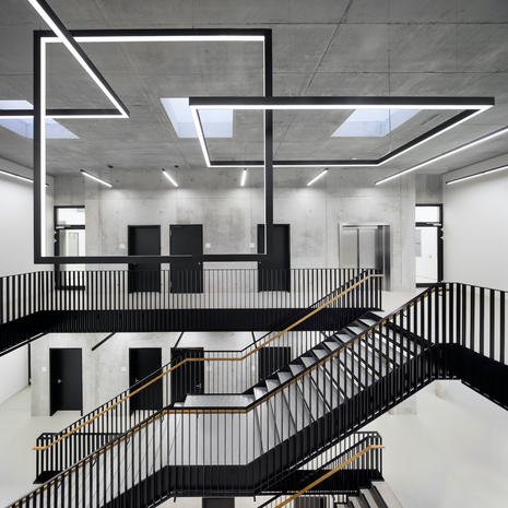 The photo shows a large, modern stairwell with clean lines and a striking LED light installation on the ceiling. Exposed concrete, black railings, and light-colored floors create an industrial, minimalist look.