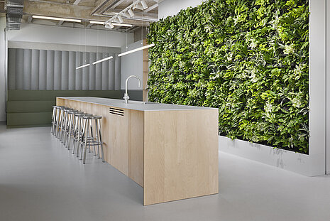 The lounge and community area feels bright and inviting, with a lush green wall adding a strong natural touch. Simple, modern stools are placed at the counter. The atmosphere is open, friendly, and inviting.