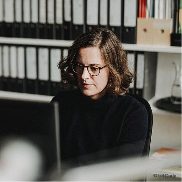 The woman is working intently at her desk in an archive or office area. The image conveys a sense of calm, focus, and authenticity.