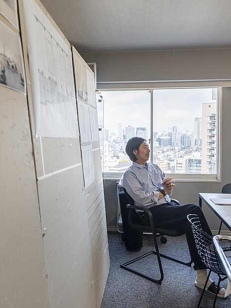 A man is sitting in an office in front of a large window overlooking a skyline. The setting feels creative, personal, and slightly improvised.