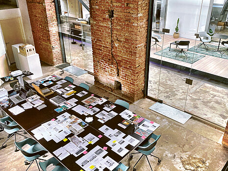 The view is from above, showing a long black table in an industrially styled meeting room, with several sheets of paper bearing notes and Post-its scattered across it. Surrounding the table are Occo chairs with black seat shells and light blue seat and back upholstery.