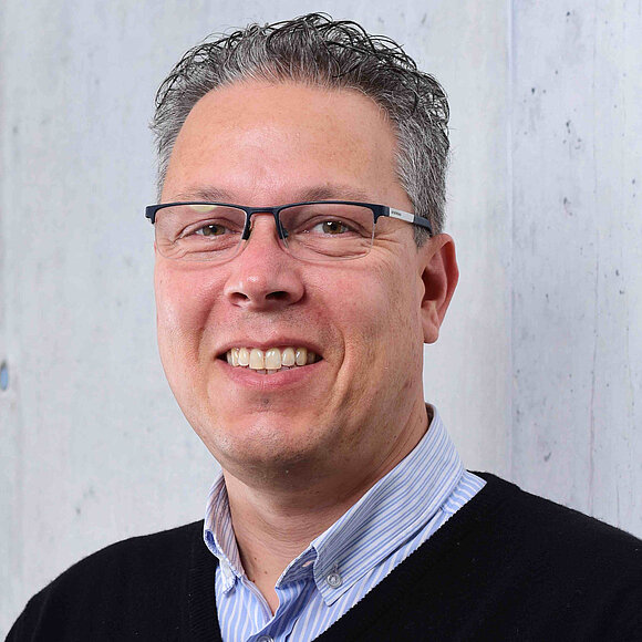 It is a portrait of a man with gray-black hair. He is wearing glasses and a black sweater.
