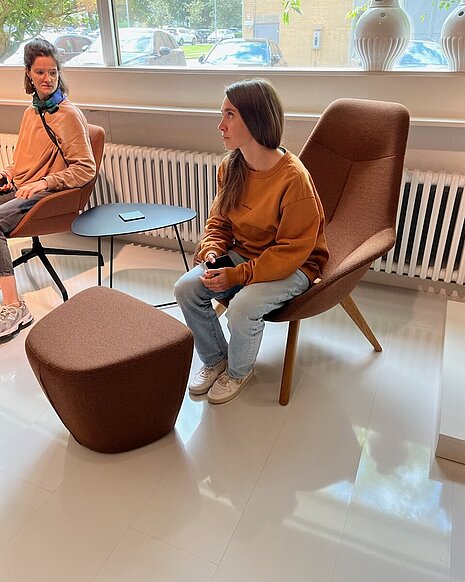 The two women are sitting in comfortable Yonda lounge chairs in a warm brown shade. The organic shapes and wooden frame create a cozy, relaxed atmosphere. The scene depicts a casual conversation in a modern lounge area.