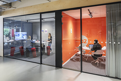 The modern office area features two glass-walled meeting rooms where several people are working. The room on the left contains several red Wilkhahn Stand-up chairs, which allow for a flexible sitting posture that encourages movement. The meeting room on the right is decorated in a bold orange and furnished with white Occo conference chairs.