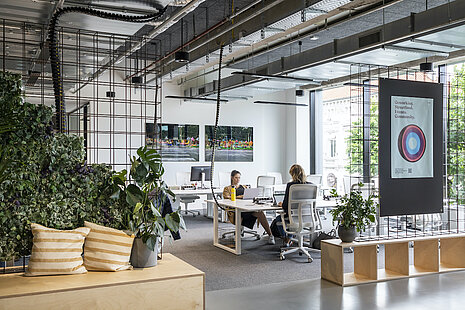 The open workspace combines an industrial look with plenty of greenery and an open floor plan. The office chairs are AT Mesh swivel chairs. The design emphasizes modern work practices, openness, and a team-oriented culture.