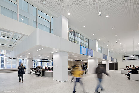 It is a large, modern hall decorated in white. On the left, people are sitting in groups of chairs, and on the right, an office area can be seen in the background.