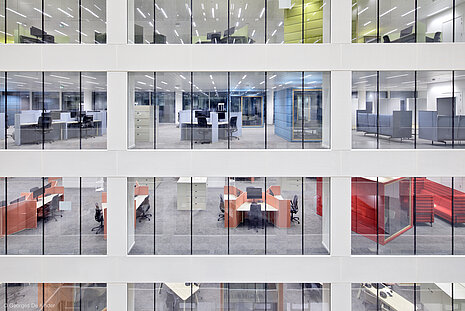 A close-up view of a building. Through the glass front, you can see the individual offices with partition walls in various colors and a few AT swivel chairs.