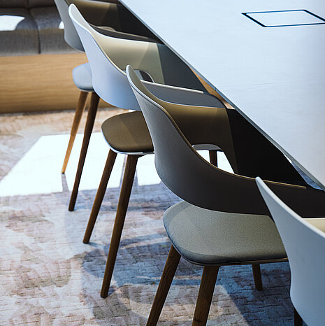 Occo chairs with white or gray seat shells and seat pads in shades of green are arranged around a white table. Die Stühle haben ein Vierfußgestell aus Holz.