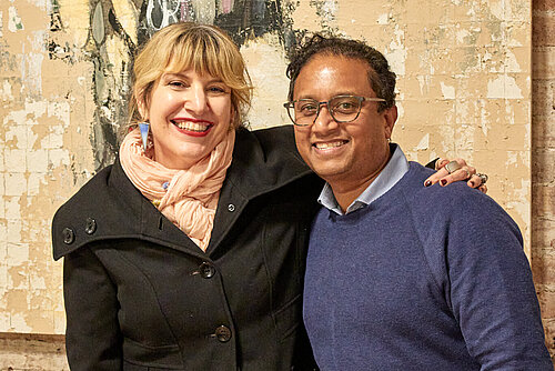It is a photo of two people standing in front of a brown brick wall. Behind them hangs an abstract painting. Both are smiling at the camera.