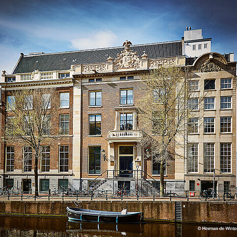 This is an exterior view of a building with a brick facade. There are many bicycles parked at the building's entrance. In front of it, you can see a river with a boat on it.