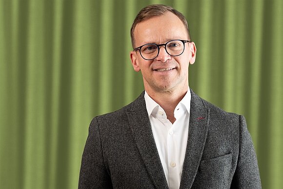 Es ist ein Bild von unserem aktuellen Geschäftsführer Götz Stamm. Er steht vor einem grünen Vorhang.