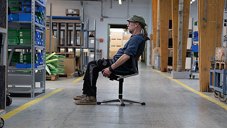 A production worker is sitting in a black Modus swivel chair located in the middle of the production hall at Wilkhahn.