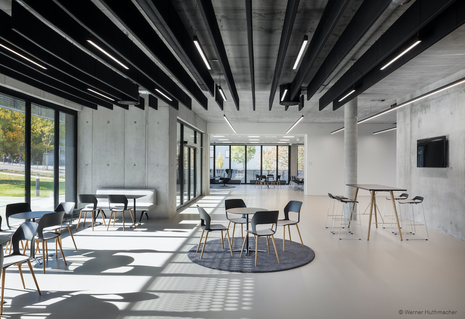 The bright interior features an open lounge and break area with various seating arrangements and tall bar tables. The black shell chairs with wooden legs closely resemble the Occo chairs, which are frequently used in modern communication areas. The space feels inviting and versatile.
