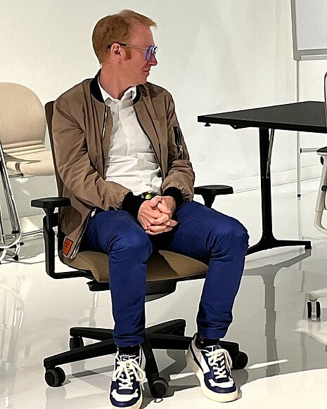 The man is sitting on an AT chair with a black frame and brown upholstery. The neutral setting highlights the chair and its comfort features. The scene looks as though he is testing or inspecting the chair's comfort.