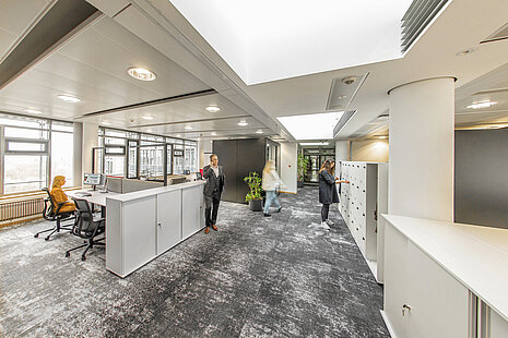 An office with white shelves on the right and a table island on the left. Black AT swivel chairs are placed around the table island.