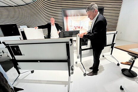 The two people are demonstrating Timetable folding tables, which can be easily unlocked and folded up to save space. The focus is on the sturdy mechanism and the mobile frames. The modern showroom highlights the functionality of the solution.
