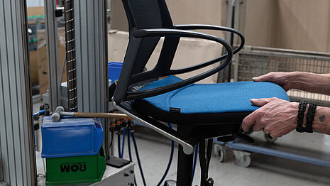 A close-up of the Modus chair being installed. The chair has a black frame and a blue seat pad.