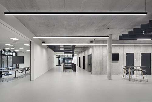 The spacious hallway, featuring exposed concrete, subdued lighting, and a central staircase, has a modern industrial feel. To the right is a tall bar table with Occo bar stools. To the left, you can see a conference room furnished with black Aline chairs.