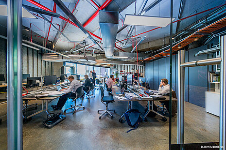 In a large office, several people can be seen going about their work. There are a few black FS swivel chairs in the room. In the background is a large window. Up near the overhead, you can see pipes and cables.