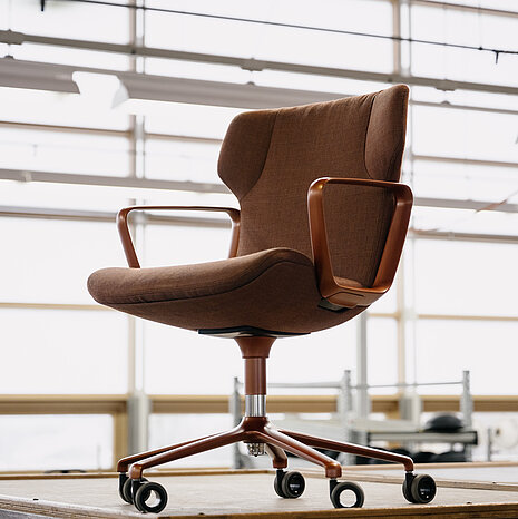 The brown-upholstered Intra swivel chair sits at a workbench in a workshop. The brown leather and color-coordinated armrests highlight the high-quality craftsmanship.