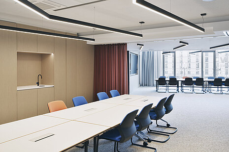 The conference room features a row of Metrik cantilever chairs in blue and orange, which stand out for their organic shape and comfortable upholstery. The flexible table arrangements and warm materials create a modern, professional meeting environment. Large windows provide plenty of natural light.