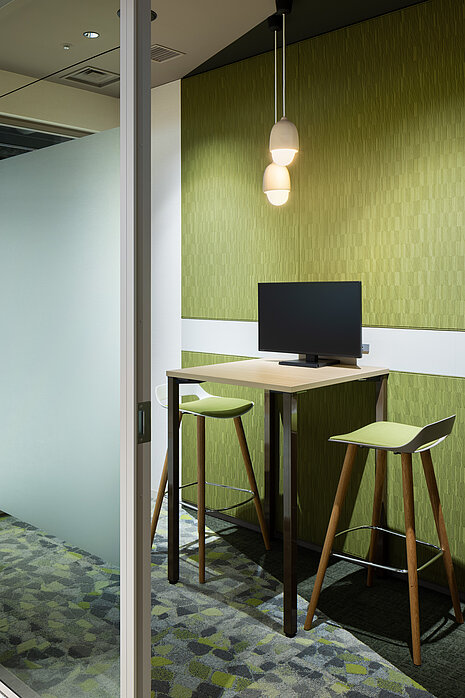 Occo bar chairs stand around a small table with a monitor on it. The setup is positioned in front of a green wall. The bar chairs have white seat shells, green seat upholstery and four-legged wooden frames.